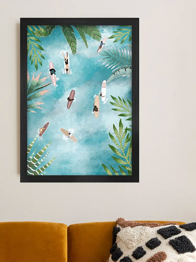 Framed artwork of people on surfboards with palm leaves against a blue sky, hanging above a sofa.
