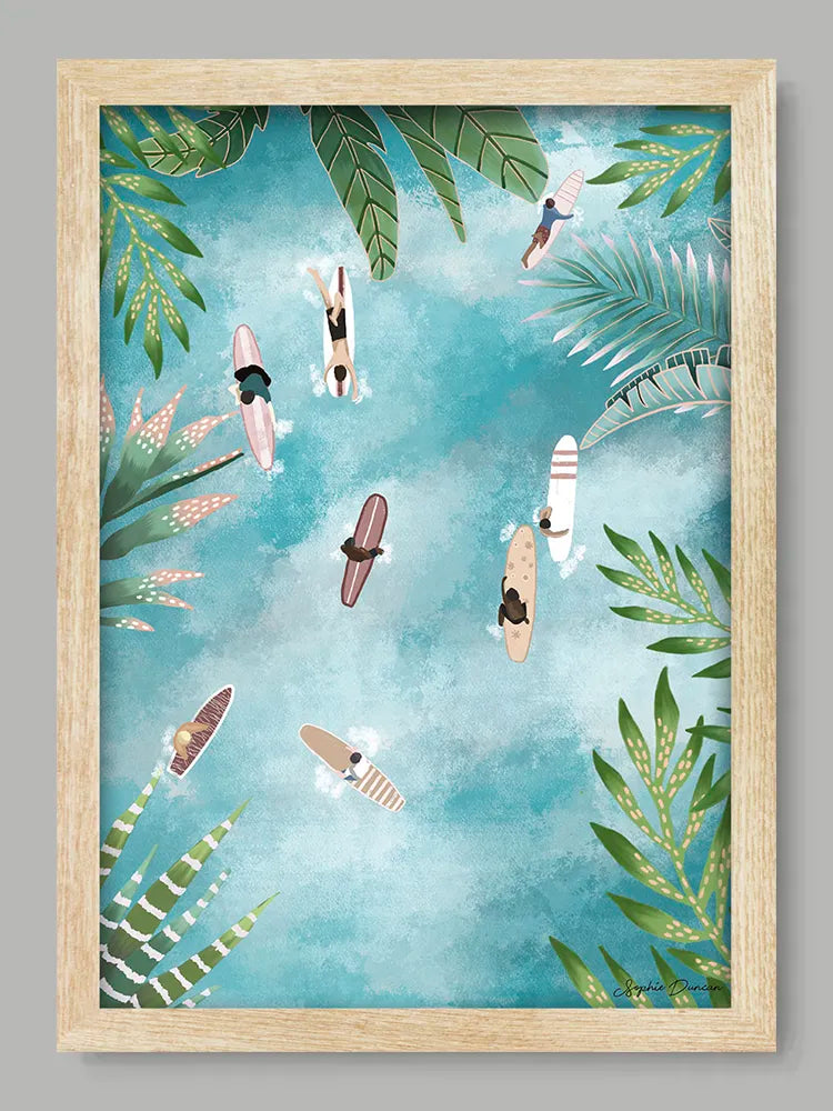Framed artwork with people on surfboards and tropical leaves against a blue sky.