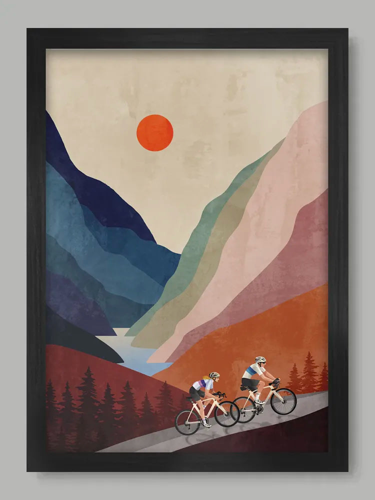 Framed artwork of two cyclists on a mountain road with a red sun and blue mountains.