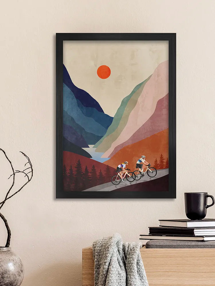 Framed artwork of cyclists on a mountain path with a red sun above, displayed on a wall.