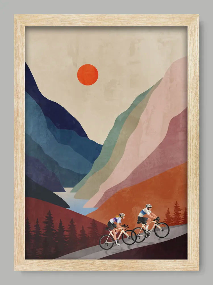 Framed artwork of two cyclists on a mountain path with a stylized mountain landscape and red sun.