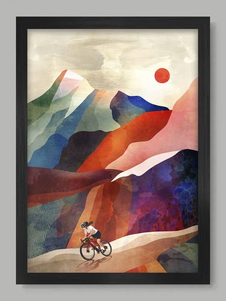 Framed artwork of a cyclist on a mountain path with colorful mountains and a red sun.