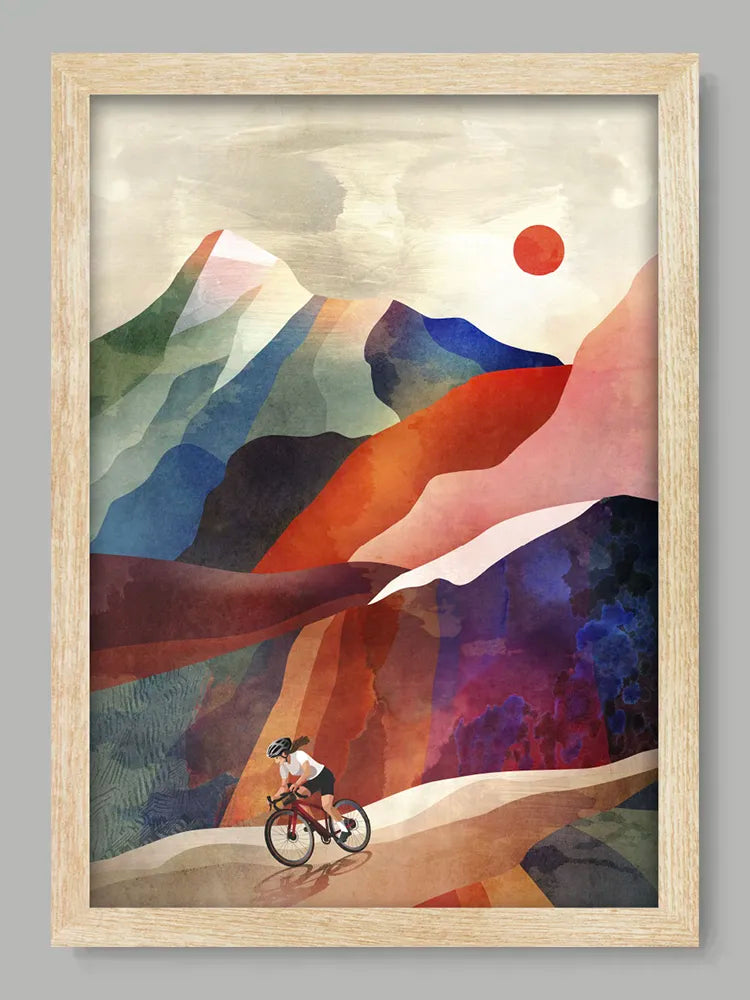 Framed artwork of a cyclist on a mountain path with colorful mountains and a sun.