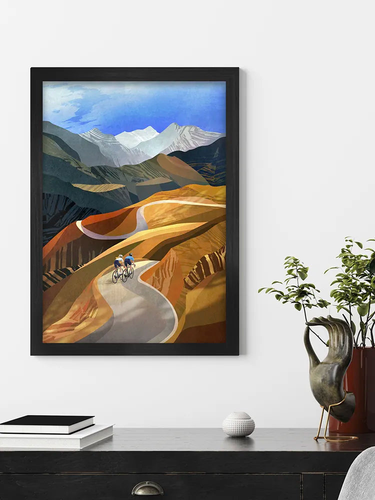 Framed artwork of a mountain landscape with a road and cyclists on a white wall.