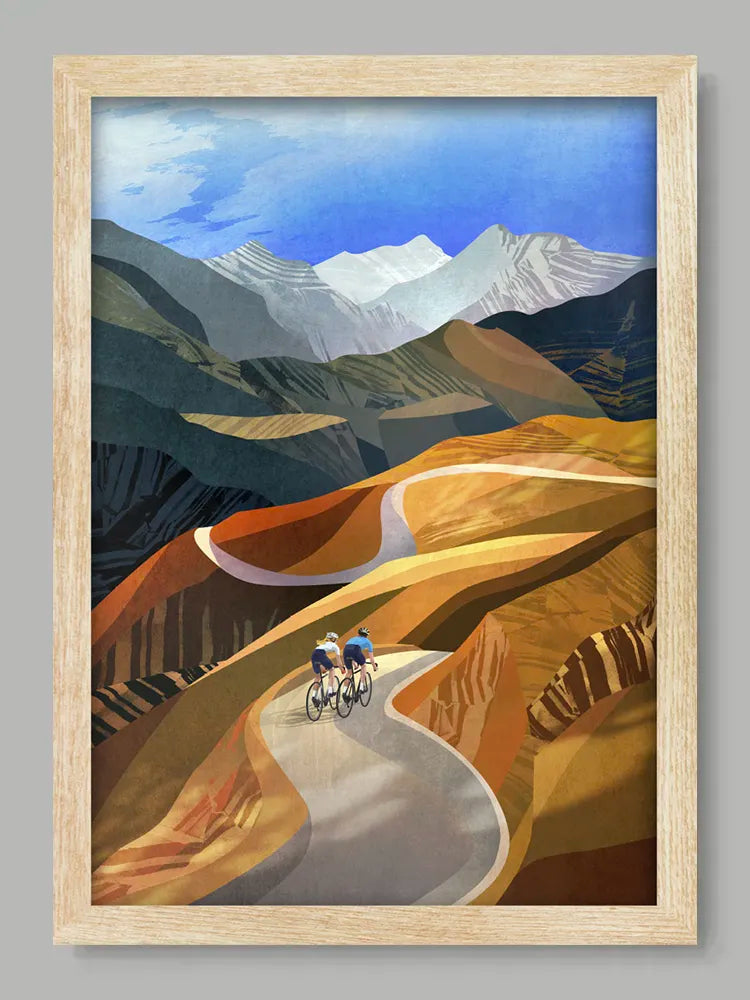 Framed artwork of two cyclists on a mountain road with mountains in the background.