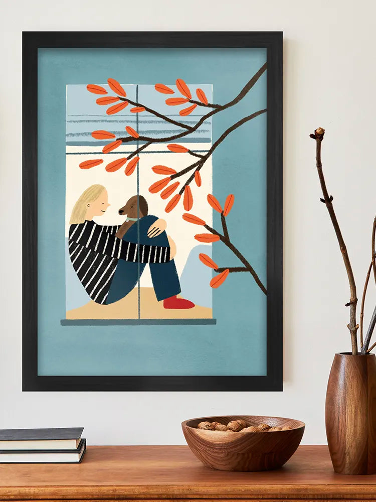 Framed artwork of two people embracing by a window with red leaves on a wall above a wooden shelf.